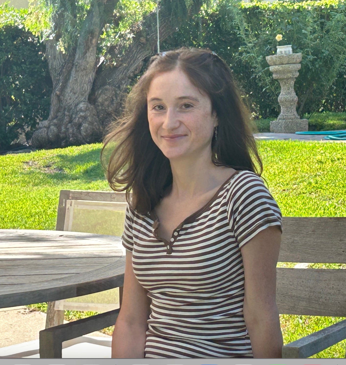 Adele sits at a wooden bench and table, wearing a striped shirt and smiling at the viewer.